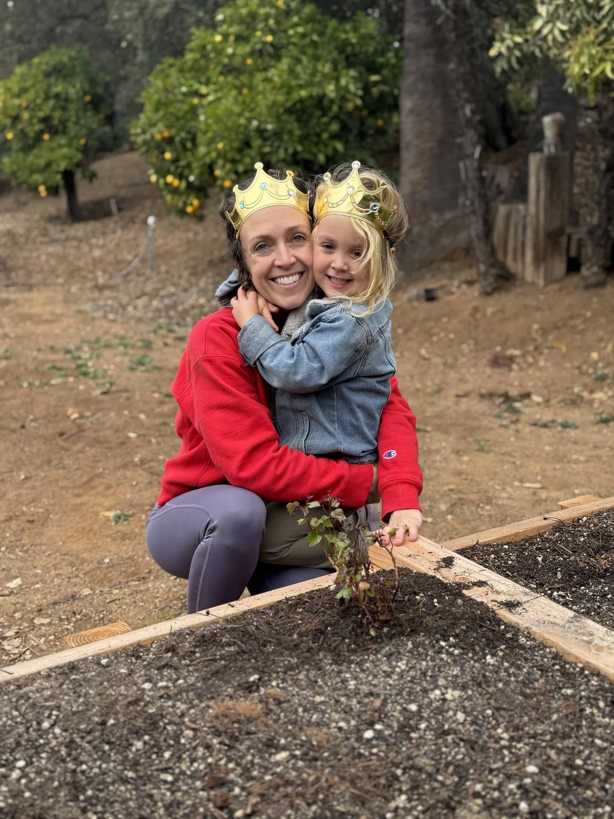 A mother and a three-year-old toddler kneeling in the dirt, carefully planting a sprouted sweet potato into a raised bed