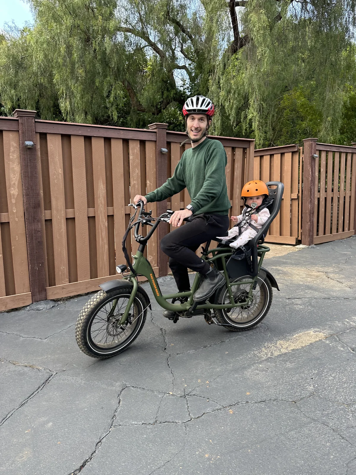 A dad in a bike helmet astride an olive-green electric cargo bike, with a baby in an orange helmet and pacifier strapped into the child seat on the back, in front of a wooden fence