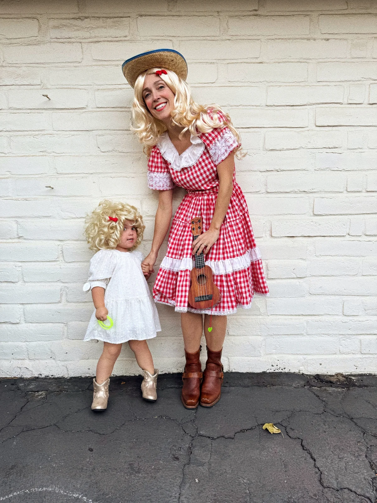 Mother and toddler daughter dressed as Dolly Parton in blonde wigs — one in a red gingham dress holding a toy ukulele, the other in a white dress and gold cowboy boots — in front of a white brick wall
